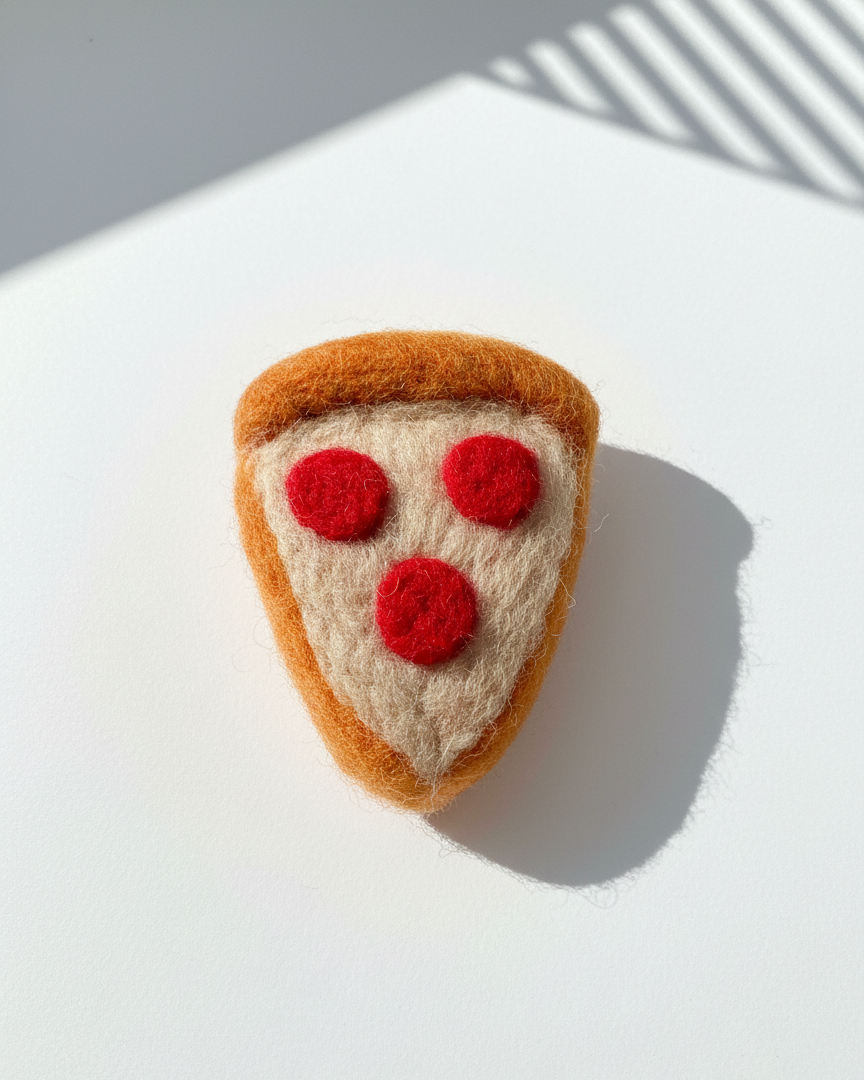 Handmade wool pizza slice fridge magnet with red felt pepperoni details on white surface