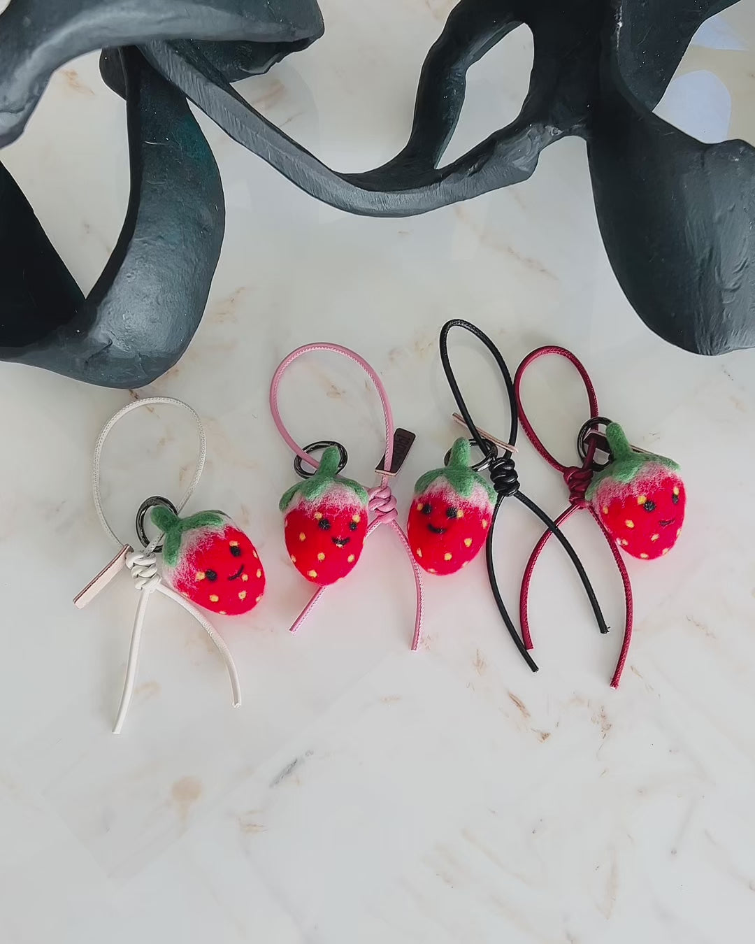 Panning video showing the full color collection of needle-felted strawberry bag charms with four leather strap options: white, pink, black, and red, arranged on a marble surface.