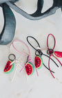 Panning video showing the full collection of needle-felted watermelon bag charms with four leather strap options: white, pink, black, and red, arranged on a marble surface.