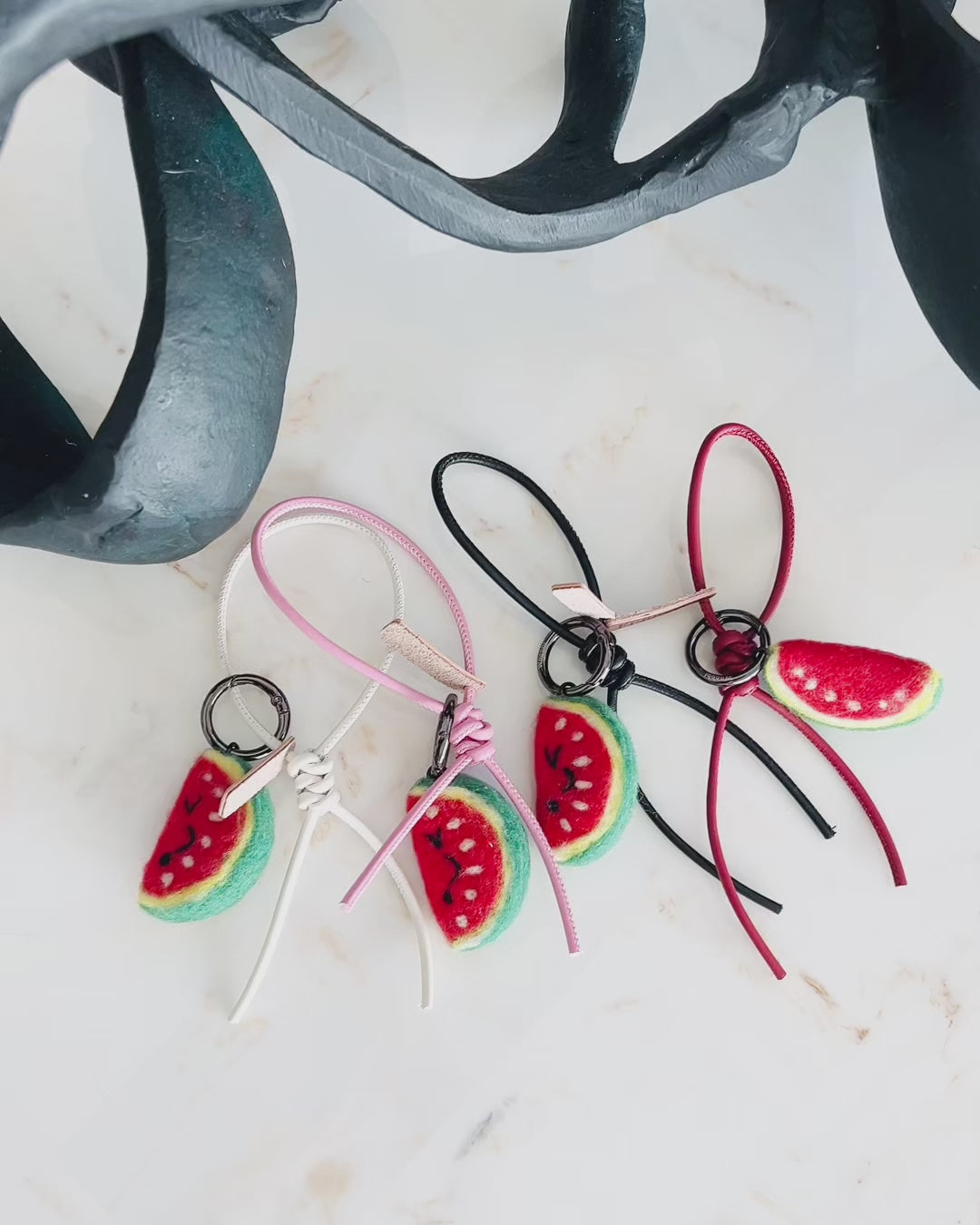 Panning video showing the full collection of needle-felted watermelon bag charms with four leather strap options: white, pink, black, and red, arranged on a marble surface.