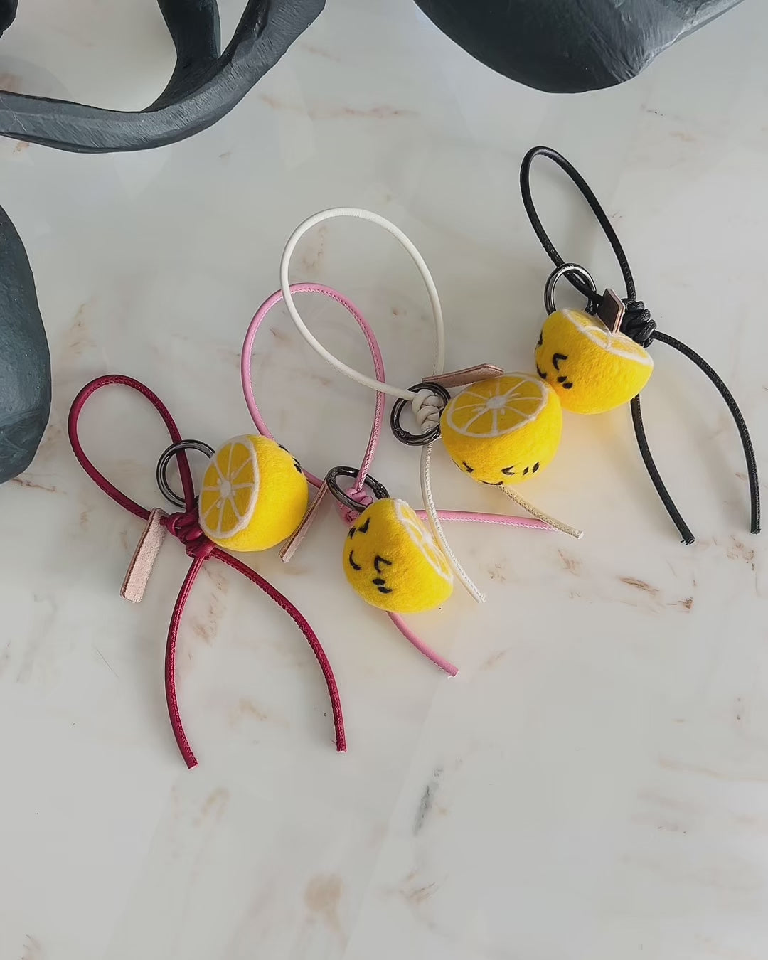 Panning video showing the full collection of needle-felted lemon bag charms with four leather strap options: red, pink, white, and black, arranged on a marble surface.