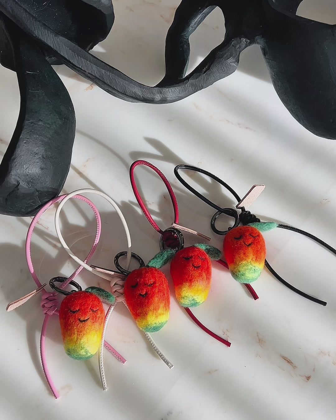 Panning video showing the full collection of needle-felted mango bag charms with four leather strap options: white, black, red, and pink, arranged on a marble surface.