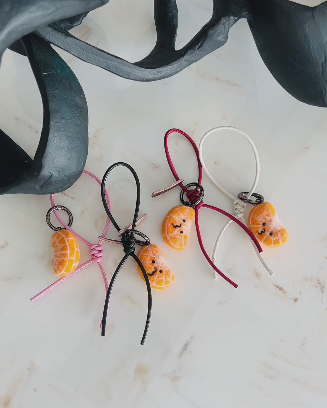 Panning video showing the full color collection of needle-felted orange slice bag charms with four leather strap options: white, pink, black, and red, arranged on a marble surface.