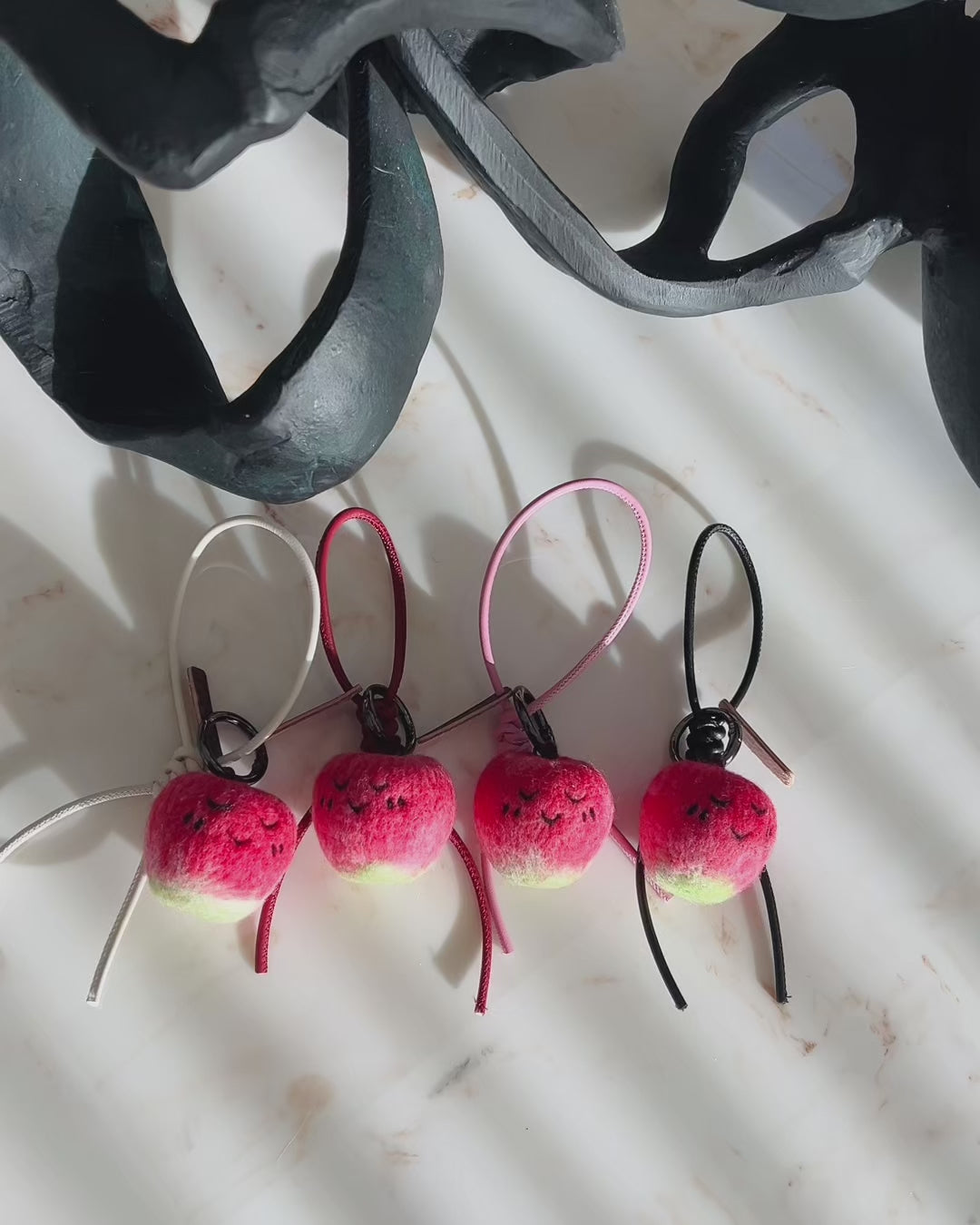Panning video showing the full collection of needle-felted apple bag charms with four leather strap options: white, black, red, and pink, arranged on a marble surface.
