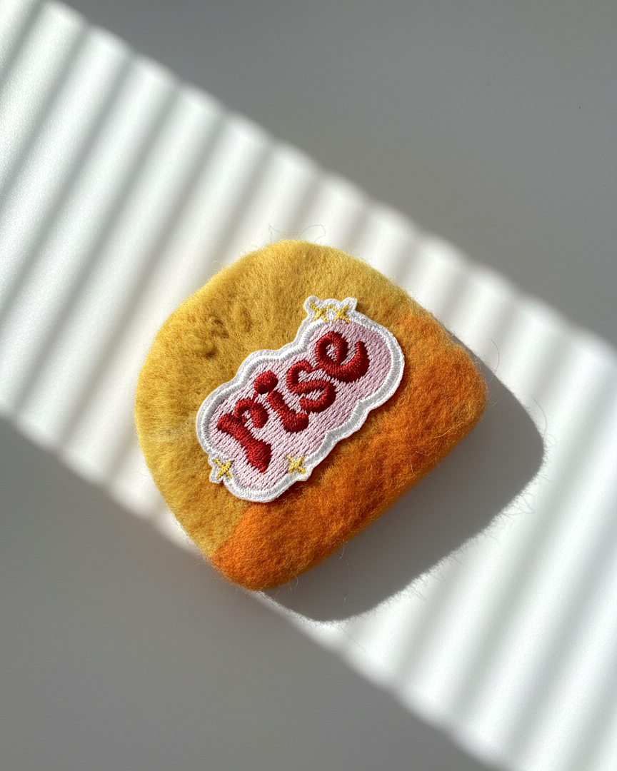Handmade wool fridge magnet in yellow and orange tones with embroidered Rise patch, needle-felted merino wool